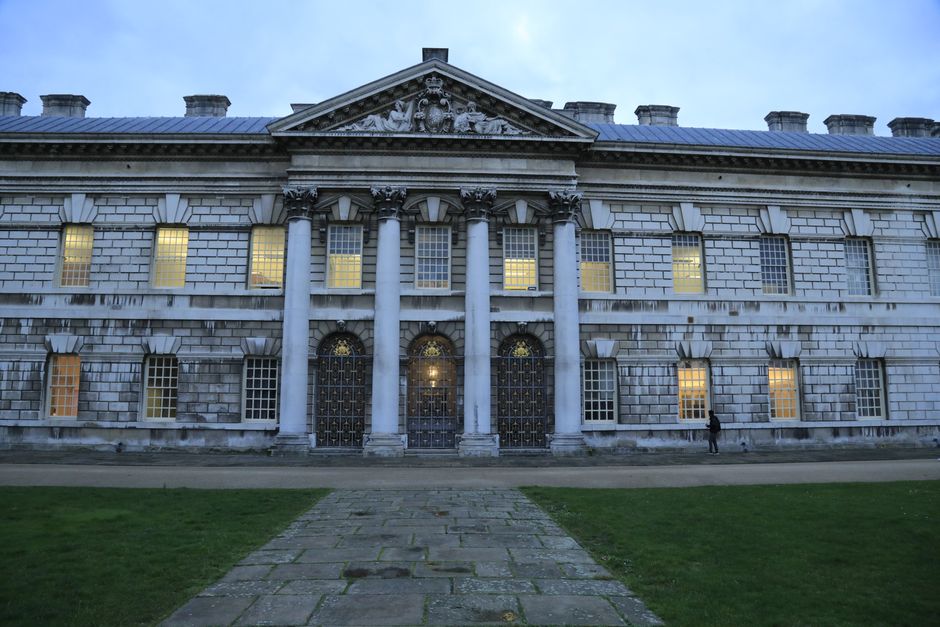 An exterior view of an ornate, historic building with classical architectural features, including a triangular pediment with decorative sculptural details, tall white columns supporting the entrance, and large rectangular windows with grid patterns. There are multiple windows illuminated from inside, suggesting interior lighting. The building's façade is constructed from light-colored stone or brick, with visible stonework detail and a slate roof. In front of the building, a paved pedestrian pathway leads directly to the entrance, which is framed by the columns. A person is seen walking along the pavement near the right side of the building. The surrounding area includes a grassy lawn, and the sky above is cloudy, indicating late afternoon or early evening. This setting could be associated with professional building management, historical preservation, or a notable public structure, relevant to house removals and relocation services by Movers Greenwich, situated within the context of packing, moving logistics, and furniture transport.