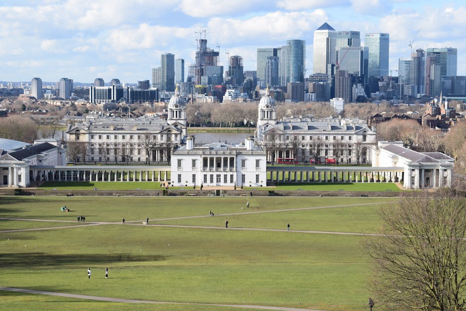 A wide view of the Royal Pavilion in Brighton, featuring its distinctive white neoclassical and oriental-inspired architecture with domed roofs and ornamental details, situated behind a large open green park area with walking pedestrians. In the background, a modern city skyline with high-rise office buildings and skyscrapers is visible under a partly cloudy sky. The park's grassy area is dotted with small groups of people, some walking and others sitting or relaxing, with paved pathways crossing the lawn. The foreground includes a leafless tree on the right edge. The scene is captured in natural daylight, highlighting the contrast between historical architecture and contemporary urban development, suitable for illustrating house or property removals, packing, and home relocation services in an urban environment.