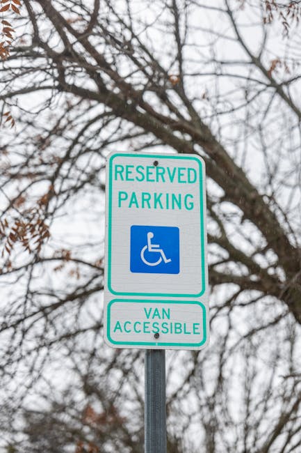 A close-up view of a green and white reserved parking sign with a blue wheelchair symbol indicating wheelchair accessibility and van access, mounted on a metal pole against a background of leafless tree branches and an overcast sky. The sign is situated outdoors, likely on a street or parking area relevant to house removals and furniture transport. The sign's purpose is to designate parking spaces reserved for disabled access and van loading, which aligns with the logistics involved in home relocations. The presence of the sign reflects the importance of proper parking permits and designated loading zones for removal vans to facilitate efficient and compliant moving services, as offered by Movers Greenwich, specialists in removals and relocation logistics.