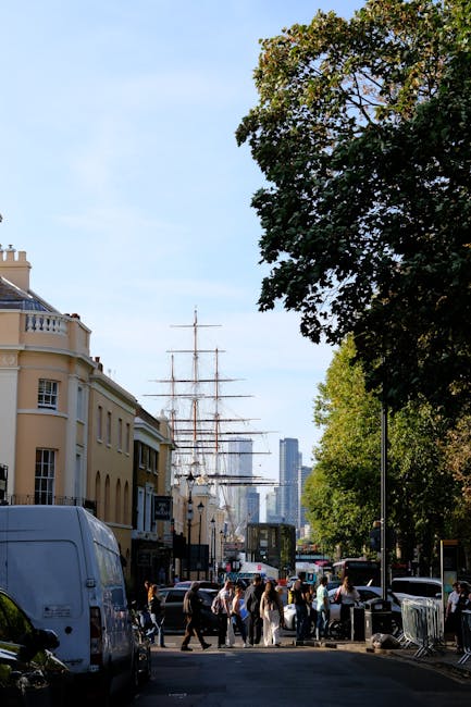 A busy street scene in Greenwich near the Cutty Sark, featuring a large ships mast and square-rigged sailing ship moored along the riverbank in the background. The foreground shows pedestrians walking