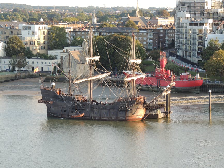 A busy street scene in Greenwich near the Cutty Sark, featuring a large ships mast and square-rigged sailing ship moored along the riverbank in the background. The foreground shows pedestrians walking