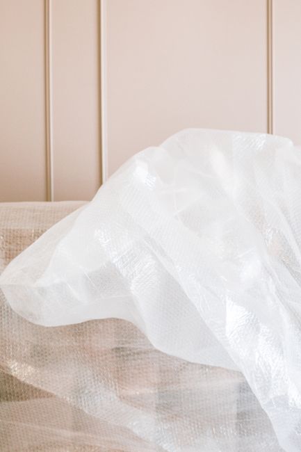 A person hand, wearing a brown T-shirt, is carefully handling a large piece of bubble wrap wrapping a piece of furniture, likely during a house removal or packing process. The furniture, possibly a ch