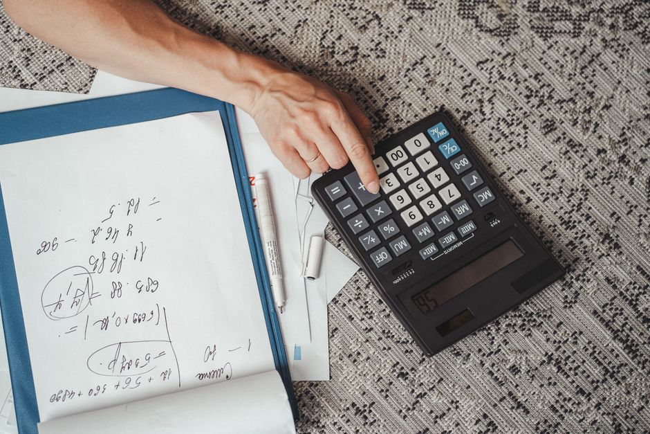 A person's hand using a small, portable green calculator with black buttons on a white surface, surrounded by a partially visible open planner or ledger, a blue and white pill bottle lying nearby, and