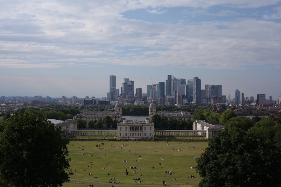 A view of a modern city skyline with numerous tall glass skyscrapers and construction cranes under a partly cloudy sky, seen from a park area with lush green trees and grass in the foreground. The sce