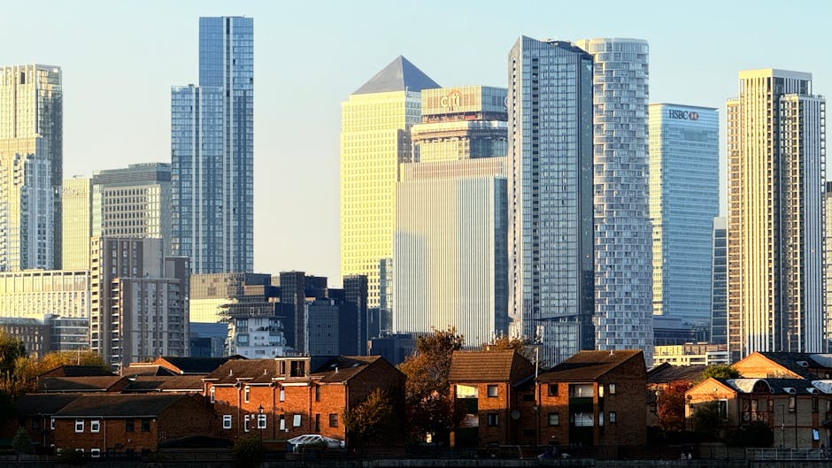 A city skyline during daylight showing a mix of modern high-rise office buildings with glass facades and older, shorter brick residential houses positioned in the foreground. The high-rise buildings feature reflective windows and prominent corporate logos, such as HSBC, in bright sunlight, highlighting their height and contemporary architectural style. The residential houses have reddish-brown brick exteriors with small front gardens, some with trees and parked cars along the street. The scene captures a typical urban environment where office relocations and furniture transport might take place, with the contrast between commercial and residential areas visible in the image. Moving equipment like trolleys or packing supplies are not visible, but the context suggests possible logistics operations occurring nearby, which local removals specialists such as Movers Greenwich could facilitate for business or home relocations.