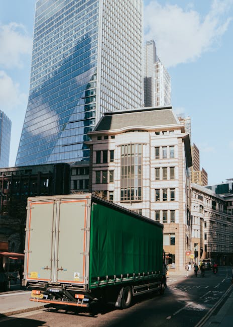 A large moving truck with a green cargo cover is parked on the street in an urban area surrounded by tall modern office buildings with glass facades and older multi-story stone structures. The truck is positioned near a sidewalk with some pedestrians walking nearby, and the vehicle appears to be in the process of a home or office relocation, likely related to furniture transport or packing and moving activities. The scene is captured during daylight hours with clear weather, and the truck's rear doors are closed, indicating it may be awaiting loading or just completed unloading as part of a professional removals service. This setting exemplifies effective logistics for house removals or office moves in central Greenwich, with the presence of equipment such as the truck and the urban environment suggesting a coordinated moving operation by Movers Greenwich.