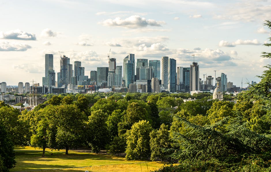 A city skyline during daylight showing a mix of modern high-rise office buildings with glass facades and older, shorter brick residential houses positioned in the foreground. The high-rise buildings feature reflective windows and prominent corporate logos, such as HSBC, in bright sunlight, highlighting their height and contemporary architectural style. The residential houses have reddish-brown brick exteriors with small front gardens, some with trees and parked cars along the street. The scene captures a typical urban environment where office relocations and furniture transport might take place, with the contrast between commercial and residential areas visible in the image. Moving equipment like trolleys or packing supplies are not visible, but the context suggests possible logistics operations occurring nearby, which local removals specialists such as Movers Greenwich could facilitate for business or home relocations.
