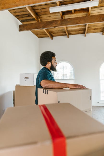 A male mover with a beard and dark hair, wearing a blue shirt, standing inside an unfinished room with a wooden ceiling and white walls, is seen leaning on a stack of cardboard boxes during a home relocation. The boxes vary in size, with some sealed using red packing tape and others open, revealing packing materials. The room features a large arched window allowing natural light to illuminate the scene. Additional cardboard boxes are stacked nearby, and there are no furnishings or furniture visible in the space. The floor is light-colored, and the overall environment appears to be in the process of packing or loading for a move, reflecting the logistics involved in furniture transport and packing and moving services by Movers Greenwich. The image captures the loading process, with the boxes positioned towards an open doorway, possibly leading to a moving van or vehicle outside.