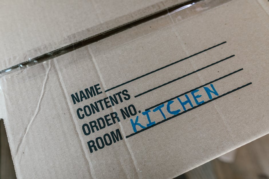 A male mover with a beard and dark hair, wearing a blue shirt, standing inside an unfinished room with a wooden ceiling and white walls, is seen leaning on a stack of cardboard boxes during a home relocation. The boxes vary in size, with some sealed using red packing tape and others open, revealing packing materials. The room features a large arched window allowing natural light to illuminate the scene. Additional cardboard boxes are stacked nearby, and there are no furnishings or furniture visible in the space. The floor is light-colored, and the overall environment appears to be in the process of packing or loading for a move, reflecting the logistics involved in furniture transport and packing and moving services by Movers Greenwich. The image captures the loading process, with the boxes positioned towards an open doorway, possibly leading to a moving van or vehicle outside.