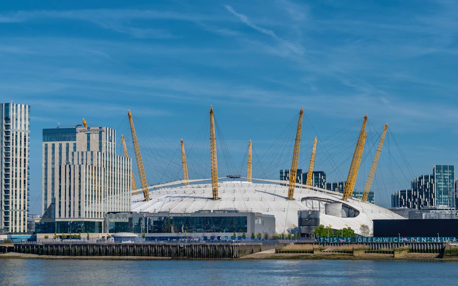 A wide-angle view of the O2 Arena in Greenwich, London, under a bright blue sky with wispy clouds. The arena's distinctive white, dome-shaped roof is supported by multiple yellow structural masts and tension cables, with adjacent modern high-rise buildings featuring glass facades visible behind it. In the foreground, a river reflects the sky and the buildings, while a promenade runs along the waterfront. This scene captures the iconic structure during daylight hours, illustrating the urban landscape of Greenwich, an area known for its vibrant skyline and waterfront location. The image is relevant to house removals and relocation services, exemplifying the area where furniture transport and home moving activities often take place, supported by companies like Movers Greenwich, who handle packing, loading, and transportation processes.